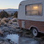 A small travel trailer parked off grid on a frosty high desert plateau at first light, with a propane cylinder standing on a wooden block beside it.