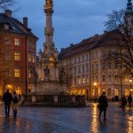 A European city square at blue hour with a baroque stone column, wet cobblestones and a tram passing at the edge of the frame.