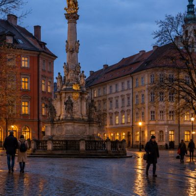 A European city square at blue hour with a baroque stone column, wet cobblestones and a tram passing at the edge of the frame.